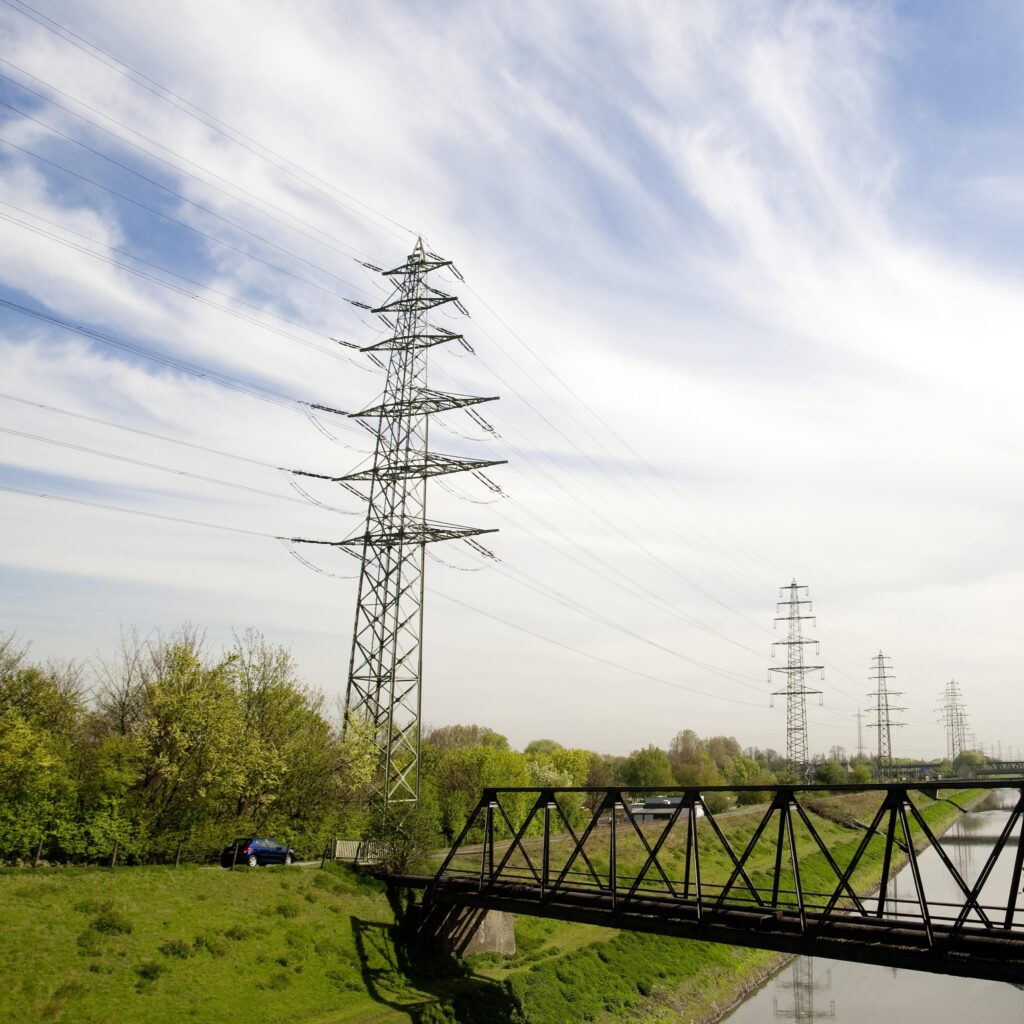 Strommasten und Hochspannungsleitungen verlaufen über eine grüne Landschaft mit Bäumen und einem Fluss. Im Vordergrund führt eine dunkle Metallbrücke über das Wasser, darüber zieht ein bewölkter, leicht streifiger Himmel vorbei.