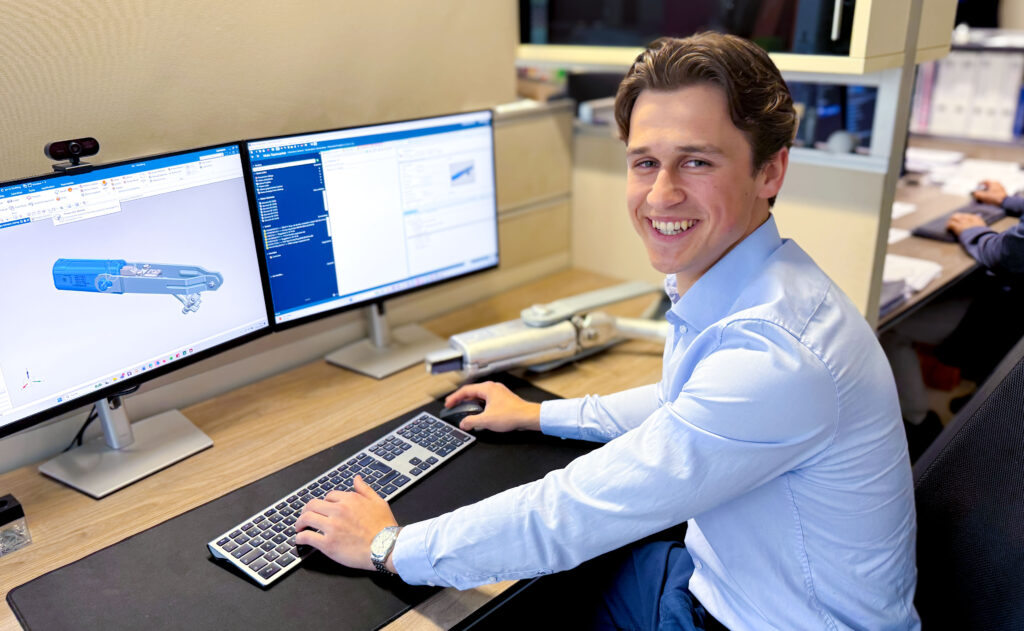 Junger Ingenieur sitzt an einem Arbeitsplatz im Büro und arbeitet an einer technischen CAD-Konstruktion auf zwei Monitoren. Auf dem Bildschirm ist ein 3D-Modell eines Bauteils für Freileitungstechnik zu sehen, während er lächelnd zur Kamera blickt und die Maus bedient.