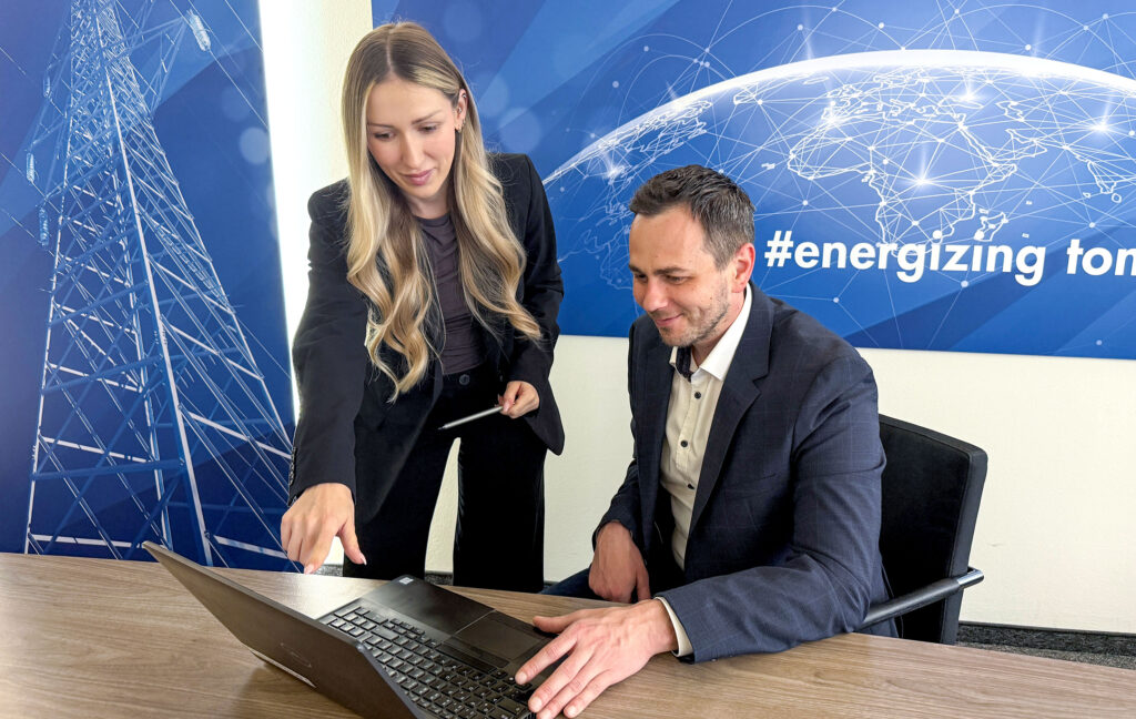 Zwei Personen arbeiten gemeinsam an einem Laptop in einem modernen Besprechungsraum; im Hintergrund sind Grafiken zur Energie‑Infrastruktur zu sehen.