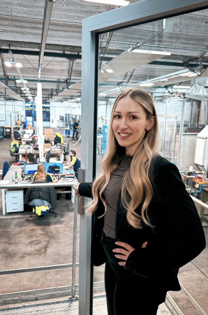 Person im Business‑Outfit steht an einer Glasbrüstung in einer Produktions‑ oder Werkshalle; im Hintergrund sind Mitarbeitende an Arbeitsplätzen zu sehen.