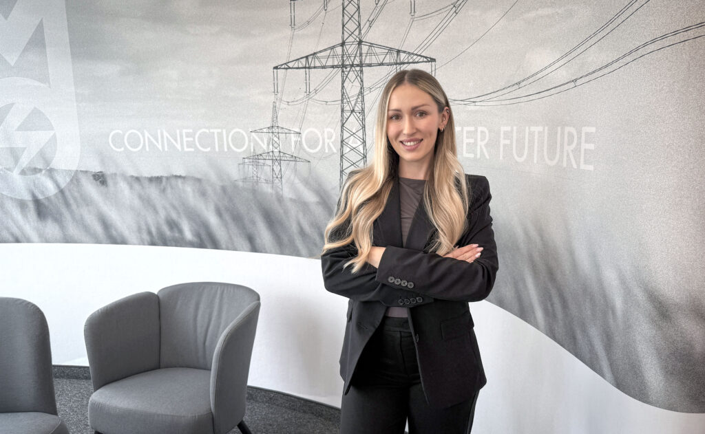 Person im Business‑Outfit steht in einem modernen Arbeitsumfeld vor einer Wandgrafik mit Strommasten und Hochspannungsleitungen.
